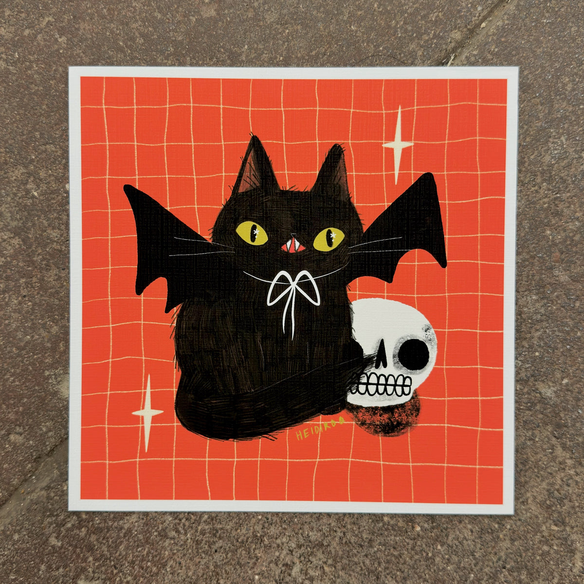 Black cat with bat wings and a white bow beside a grinning skull on a red-orange grid background, art print by Heidi Moreno on display at We Are 1976