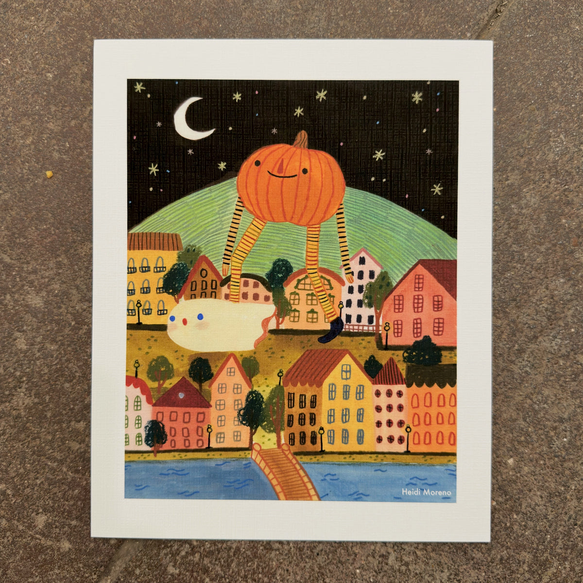 Giant smiling pumpkin on long striped legs walking over a colorful hillside town at night with a small ghost and a crescent moon, art print by Heidi Moreno on display at We Are 1976