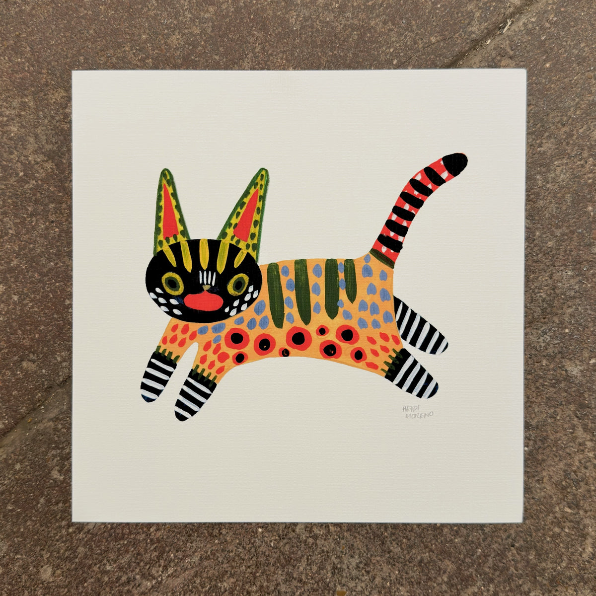 Leaping cat painted in alebrije style with a black face, spotted orange body, striped legs and a red striped tail, art print by Heidi Moreno on display at We Are 1976