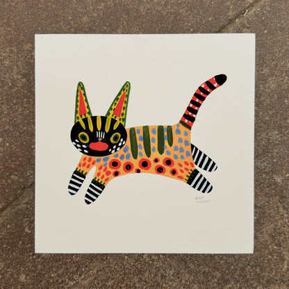 Leaping cat painted in alebrije style with a black face, spotted orange body, striped legs and a red striped tail, art print by Heidi Moreno on display at We Are 1976