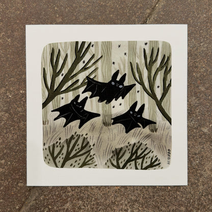 Three black bats flying through a sage-green forest of pale trees and bare branches with small stars, art print by Heidi Moreno on display at We Are 1976