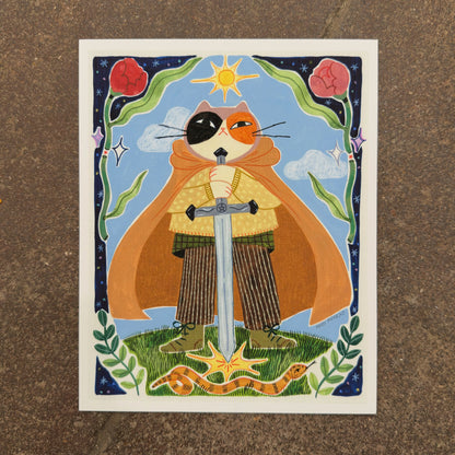 Calico cat in an orange cape holding a large sword planted in the grass with a sun above, a snake below and a border of roses and stars, art print by Heidi Moreno on display at We Are 1976