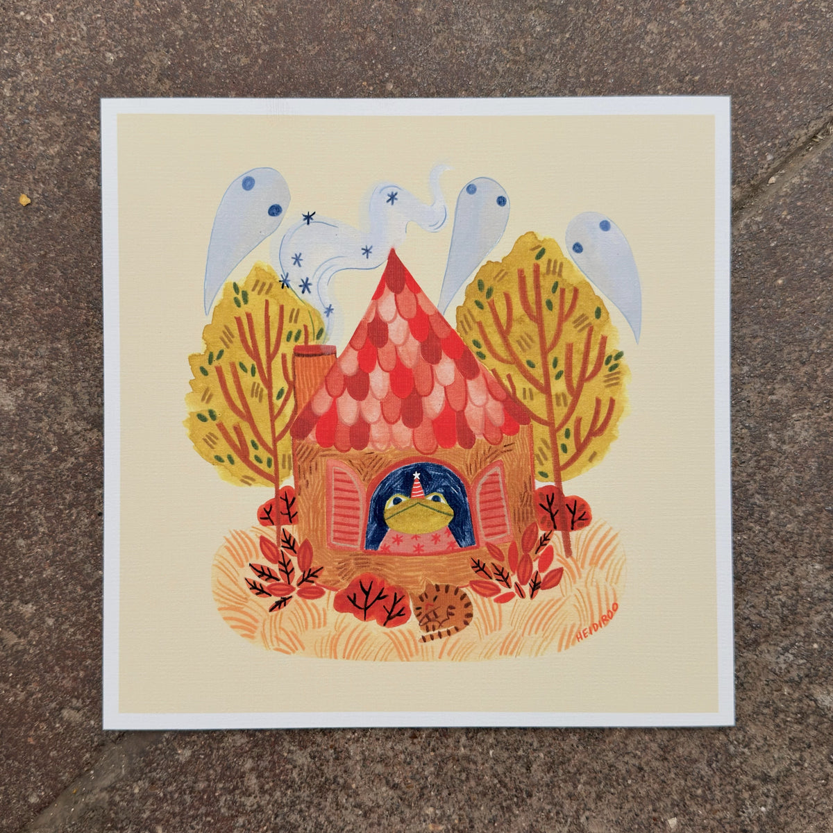 Toad in a party hat looking out the window of a red-roofed cottage with ghosts in the chimney smoke, autumn trees and a sleeping cat, art print by Heidi Moreno on display at We Are 1976