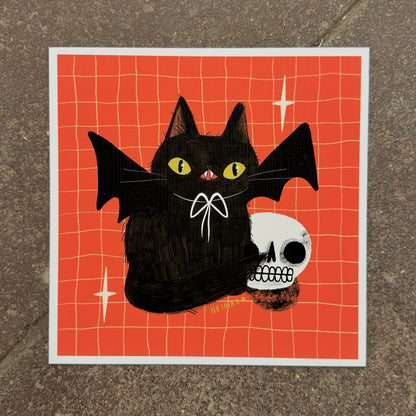 Black cat with bat wings and a white bow beside a grinning skull on a red-orange grid background, art print by Heidi Moreno on display at We Are 1976