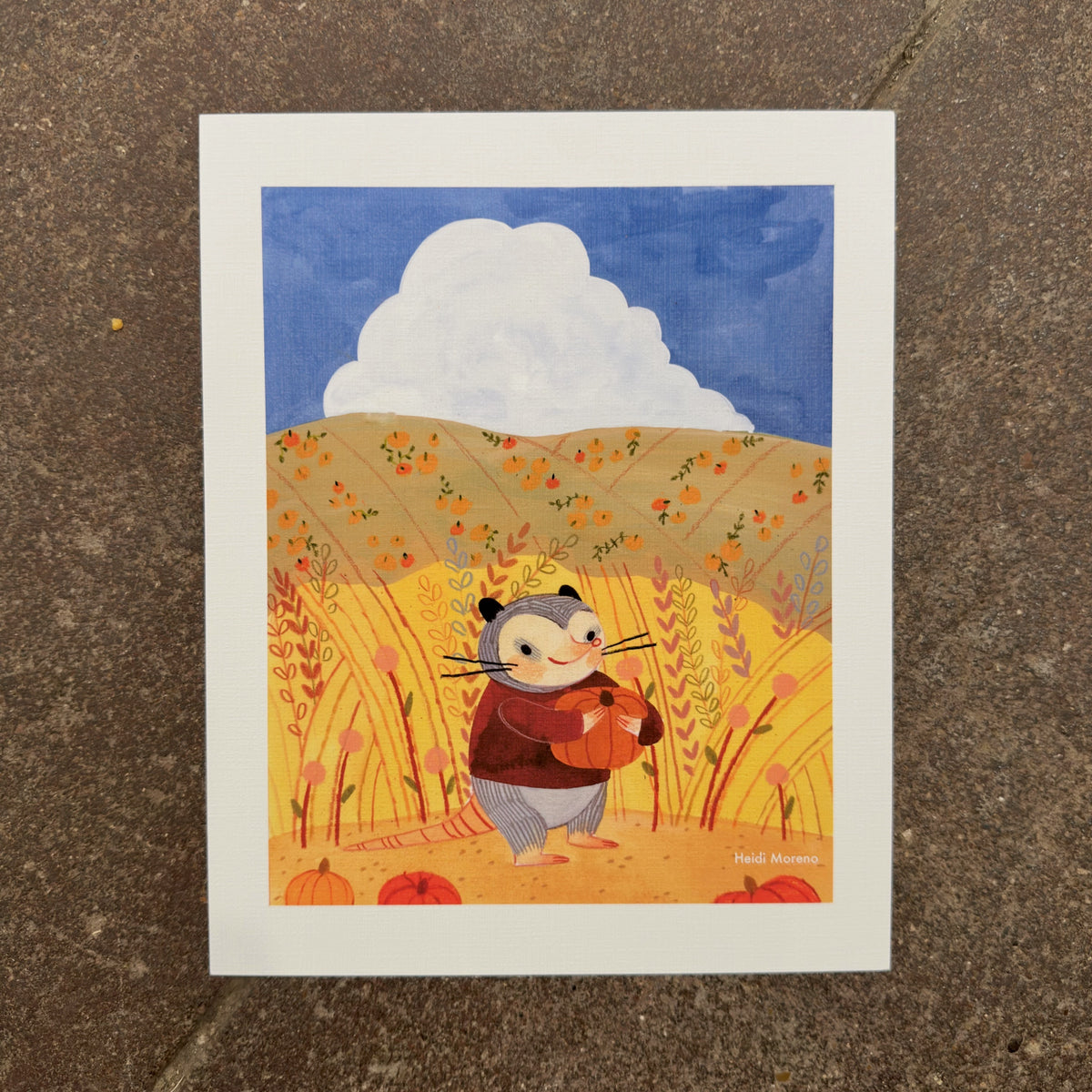 Opossum in a maroon sweater hugging a pumpkin in tall golden grass with pumpkin-dotted hills and a white cloud, art print by Heidi Moreno on display at We Are 1976