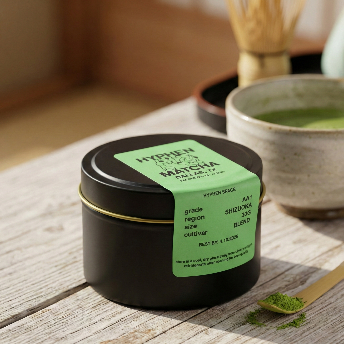 Black container of matcha powder with a green label on a wooden surface, next to a bowl of matcha and a spoon.