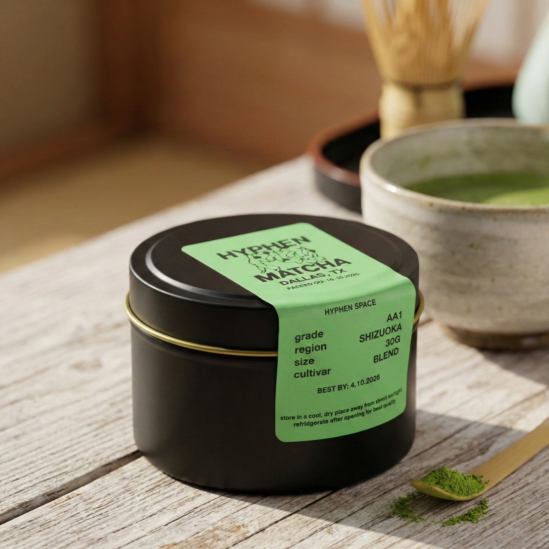 Black container of matcha powder with a green label on a wooden surface, next to a bowl of matcha and a spoon.