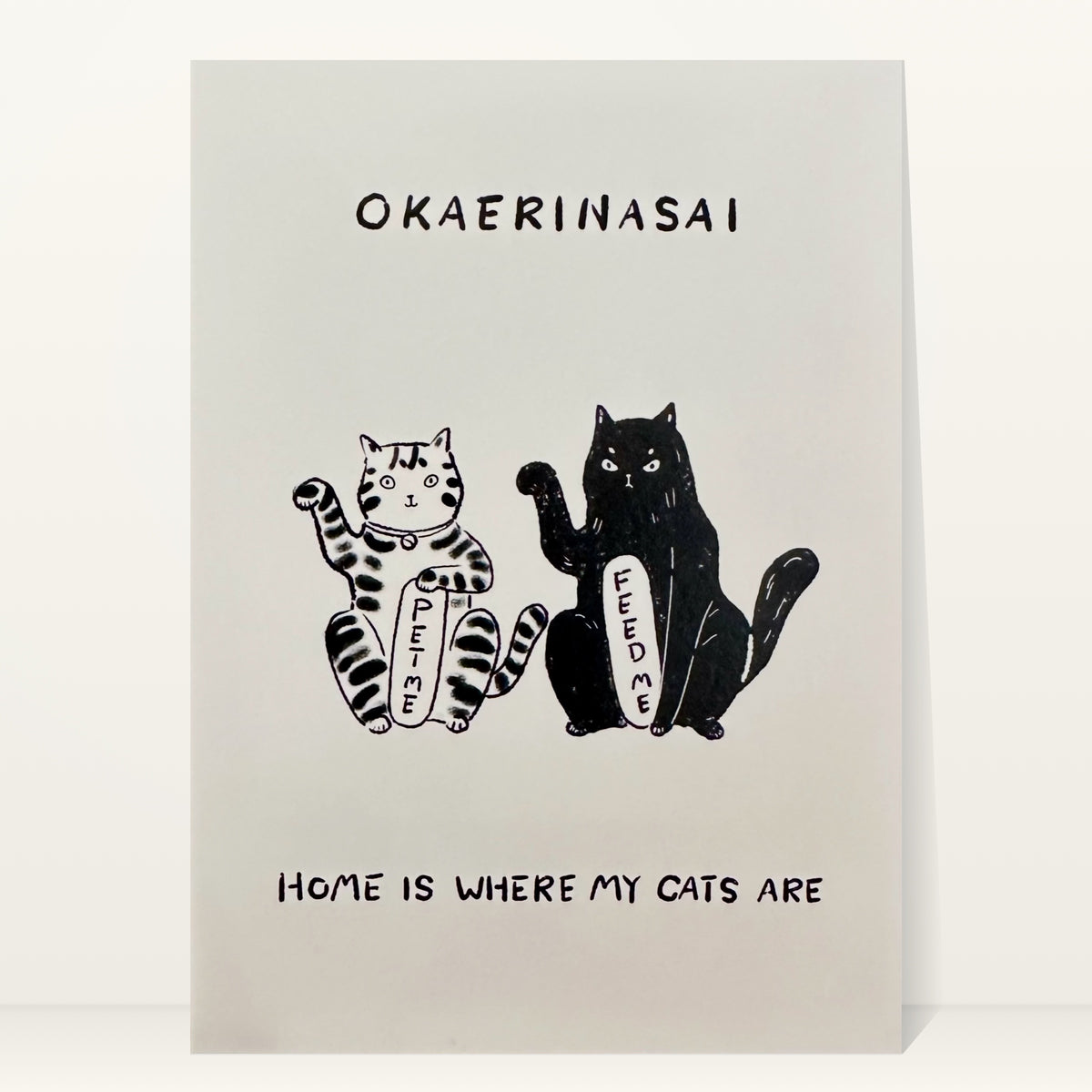 Okaerinasai mini art print by Linnan Ye: tabby cat and black cat holding “Feed me” signs, with the words “Okaerinasai” and “Home is where my cats are”