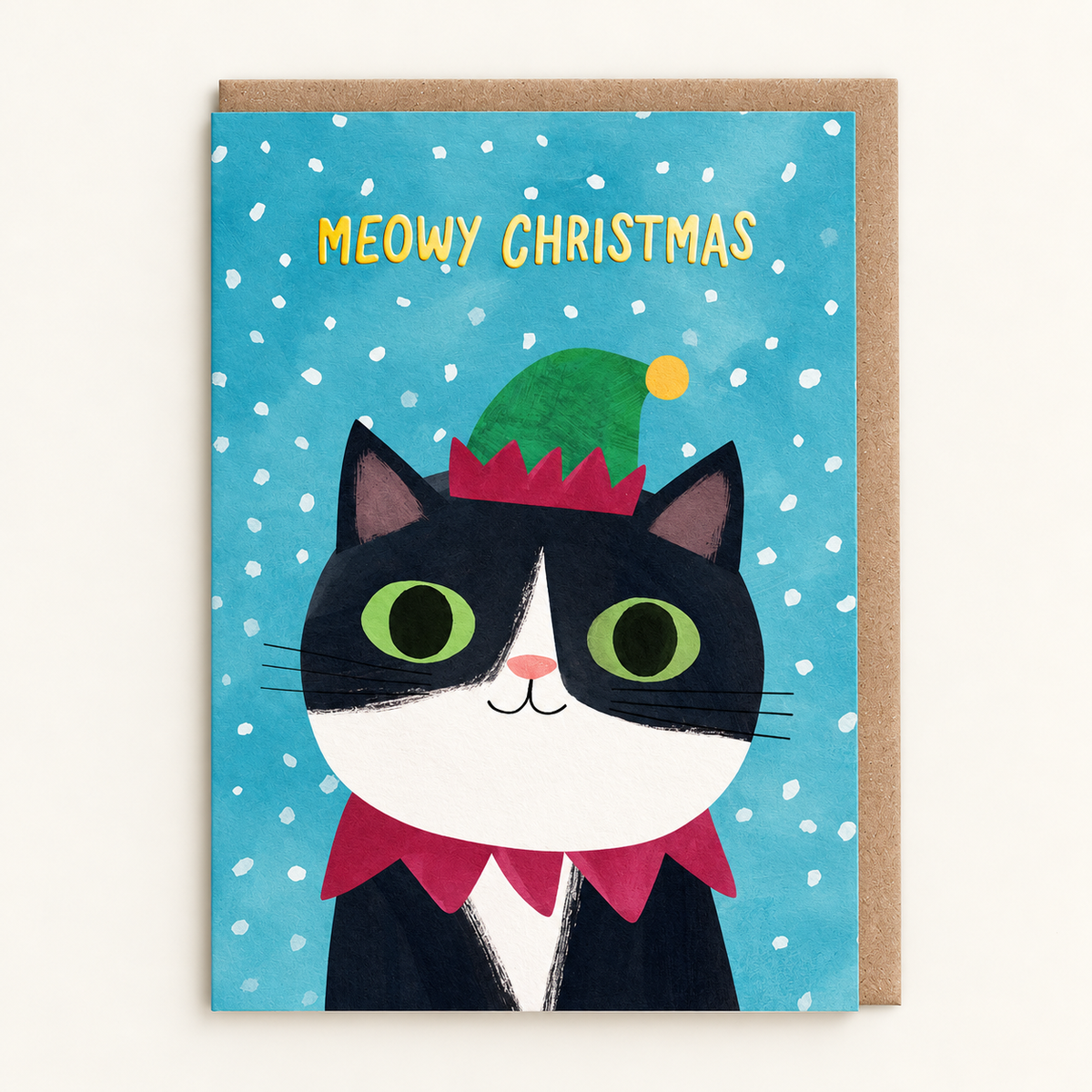 Elf Hat Kitty Christmas Card by Angela Rozelaar: painted black-and-white cat in a green elf hat and red collar under gold Meowy Christmas lettering on a snowy blue background, with a brown kraft envelope