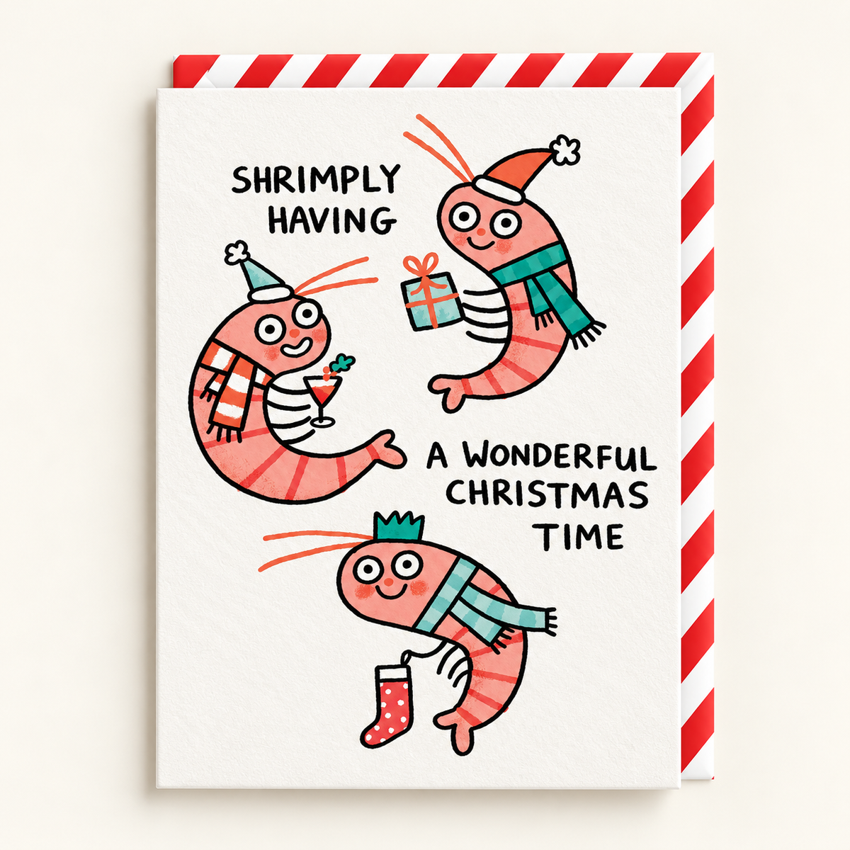 Shrimply Wonderful Christmas Card by Gemma Correll: three cartoon shrimp in hats and scarves with the words Shrimply having a wonderful Christmas time, with a red and white striped envelope